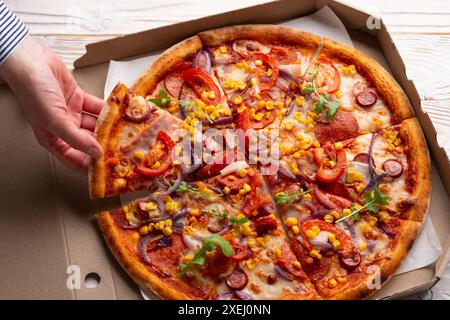 La main caucasienne humaine prend une tranche de grande pizza de la boîte de carton sur la table en bois blanc Banque D'Images