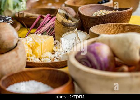 Scène rustique d'une dalle de fromage jaune dans un vieux bol en bois, sur une table parmi d'autres bols en bois de légumes Banque D'Images