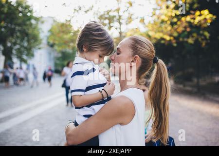 Une mère tient amoureusement son jeune fils dans ses bras, les visages rapprochés alors qu'ils partagent un moment d'intimité tranquille au milieu du parc animé de la ville. Banque D'Images