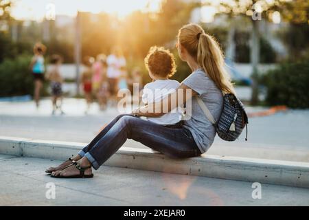 Une mère est assise avec son tout-petit sur un trottoir, regardant les autres enfants jouer dans le parc alors que le soleil se couche en arrière-plan. Banque D'Images