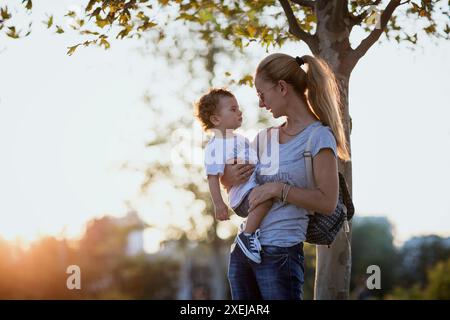 Une mère tient son petit garçon près de lui, le regardant dans les yeux. Le soleil couchant les baigne d'une lueur chaude alors qu'ils se tiennent sous un arbre avec des feuilles d'or. Banque D'Images