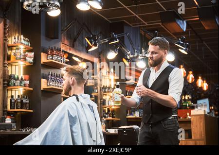 Barbier professionnel pulvérisant la lotion après-rasage sur la nuque du client. Vue arrière d'une coupe de cheveux fraîche par un coiffeur expérimenté. Coiffeur masculin utilisant un équipement spécial pour le coiffage. Banque D'Images