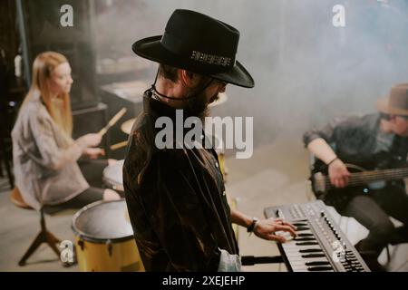 Vue latérale d'un musicien barbu portant un chapeau jouant un synthétiseur avec un groupe de rock sur scène Banque D'Images