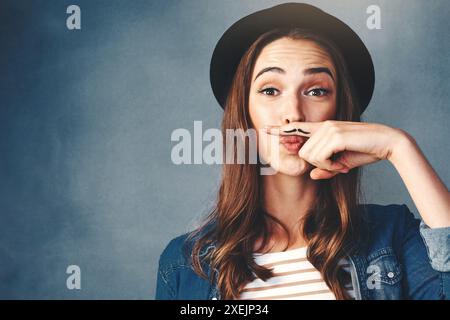 Portrait, hipster et modèle féminin avec moustache au doigt en studio pour la mode moderne, vêtements élégants ou en denim sur fond bleu. Comédie, main et Banque D'Images