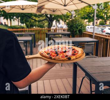 Serveur portant une pizza fraîchement cuite avec des légumes et des olives sur une assiette en bois dans une terrasse de restaurant Banque D'Images