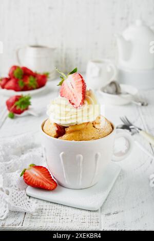 Gâteau mug fraise banane. Gâteau frais fait maison dans une tasse avec fraise, banane et crème fouettée dans une tasse de tasse en céramique rustique blanche sur une table en bois blanc Banque D'Images