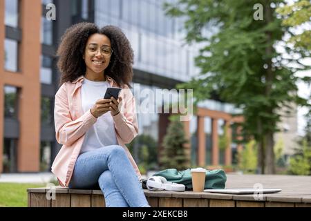Jeune étudiante assise à l'extérieur près du campus universitaire à l'aide d'un smartphone, avec tasse à café et sac à dos sur la terrasse en bois, profitant d'une journée ensoleillée, portant des vêtements décontractés et souriant Banque D'Images