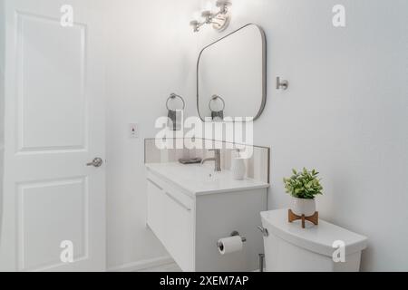 salle de bain moderne en blanc avec armoire flottante, évier, robinet, miroir, lumières, et toilettes avec des accents gris Banque D'Images