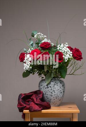 Un grand bouquet de roses rouges et de fleurs blanches dans un vase gris sur une table en bois, à côté d'une écharpe Bourgogne. Fond gris. Nature morte Banque D'Images