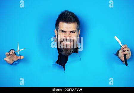 Publicité de salon de coiffure. Barbier avec rasoir droit et ciseaux regardant à travers le trou de papier. Homme barbu avec des outils de coiffure professionnels. Coiffeur s Banque D'Images