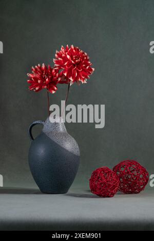 Deux dahlias rouges dans un vase gris sur fond gris, deux boules rouges se trouvent à proximité. Nature morte Banque D'Images