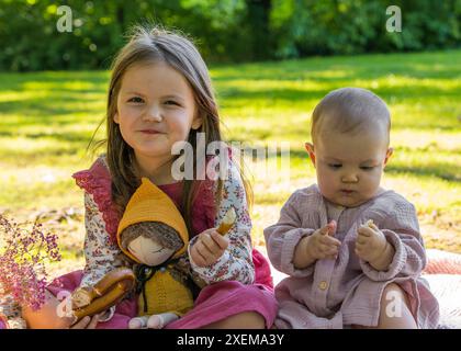 Portrait de jolies filles de 5 ans et 1 an. Deux sœurs en pique-nique. Ils ont l'air amusants devant la caméra. Il y a un ours en peluche à proximité et une application rouge Banque D'Images