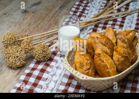 Tartes avec remplissage dans un panier en osier sur une planche de bois, à côté il y a un verre de lait et des plantes séchées. Motif rustique vue de dessus et espace de copie. Pirozh Banque D'Images