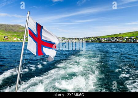 Naviguer sur le bateau à moteur de la ville de Sorvagur à l'île de Mykines sur les îles Féroé. Drapeau national des îles Féroé agitant contre un ciel nuageux. Photographie de paysage Banque D'Images