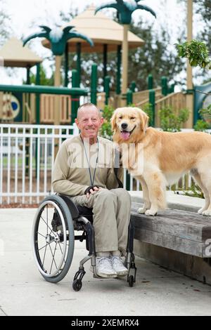 Portrait informel extérieur d'un homme en fauteuil roulant et de son chien dans une aire de jeux pour enfants ; Boynton Beach, Floride, États-Unis d'Amérique Banque D'Images