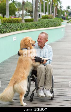 Homme en fauteuil roulant à l'extérieur jouant avec son chien d'assistance sur un chemin extérieur, l'homme et le chien se regardant face à face Banque D'Images