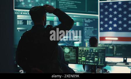 Homme militaire AMÉRICAIN supervisant l'activité de détection des menaces dans le bureau de haute technologie, examinant les données top secrètes sur grand écran. Soldats gouvernementaux gérant la guerre hybride, prévention des cyberattaques. Caméra A. Banque D'Images