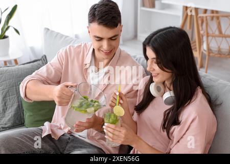 Heureux jeune couple avec des écouteurs versant du mojito savoureux dans le verre à la maison Banque D'Images