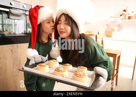 Petite fille asiatique et sa mère tenant un plateau avec des rouleaux de cannelle dans la cuisine le soir de Noël Banque D'Images