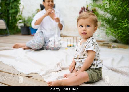 Mignon petit garçon assis sur la couverture à l'extérieur, regardant la caméra tandis que sa mère sur le fond, jouant avec lui, soufflant des bulles de savon. Joyeux carefr Banque D'Images