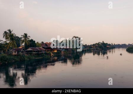 Reflets de coucher de soleil le long du fleuve Mékong avec des maisons et des palmiers bordant le rivage au Laos ; Don Khon, Province de Champasak, Laos Banque D'Images