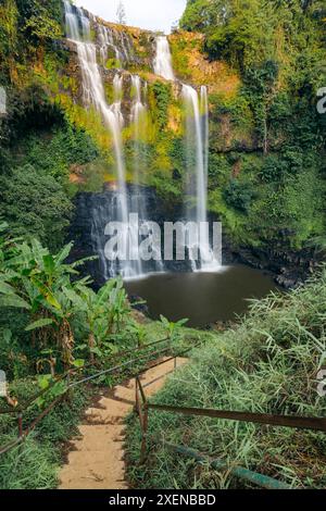 Série de cascades à Tad Gneuang Waterfall au Laos, tombant d'une haute falaise avec une végétation luxuriante et une passerelle avec garde-corps au bord de l'eau Banque D'Images