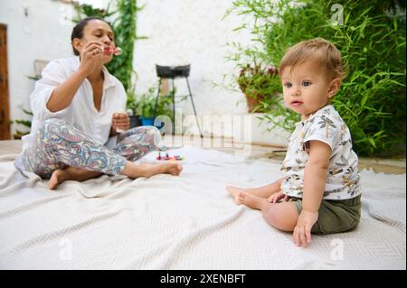 Caucasien rousse mignon tout-petit, bébé garçon attrapant des bulles de savon sur la cour, profitant de la journée d'été ensoleillée avec sa mère soufflant des bulles de savon sur le Banque D'Images