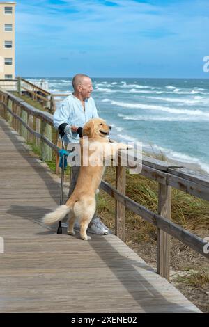 L'homme paraplégique avec des béquilles d'avant-bras et chien d'assistance apprécie la vue sur l'océan depuis une promenade, avec le chien debout contre la balustrade en bois Banque D'Images