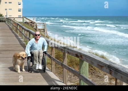 Homme paraplégique en fauteuil roulant avec chien d'assistance aime l'océan depuis une promenade ; Boynton Beach, Floride, États-Unis d'Amérique Banque D'Images