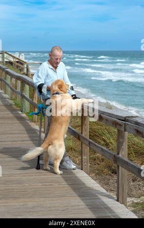 L'homme paraplégique avec des béquilles d'avant-bras et chien d'assistance apprécie la vue sur l'océan depuis une promenade, avec le chien debout contre la balustrade en bois Banque D'Images