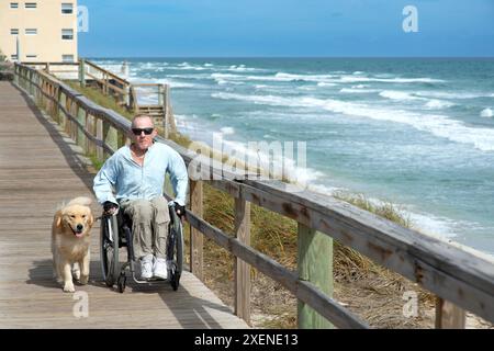 Homme paraplégique en fauteuil roulant avec chien d'assistance profite de la vue sur l'océan depuis une promenade ; Boynton Beach, Floride, États-Unis d'Amérique Banque D'Images