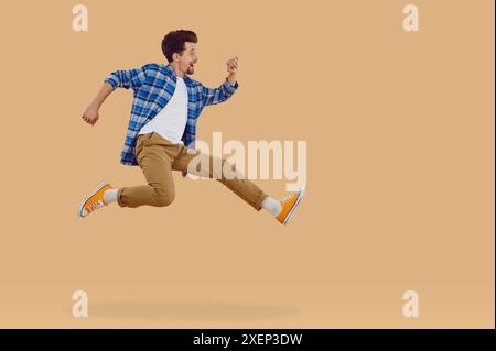 Bonne brune jeune homme dans des vêtements décontractés sautant dans l'air sur fond beige, vue de côté. Banque D'Images