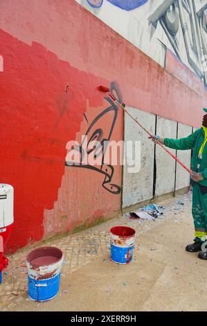 Processus d'un peintre en combinaisons vertes comme il peint sur des graffitis sur un mur rouge, couvrant de vieilles étiquettes avec de la peinture fraîche d'un rouleau Banque D'Images