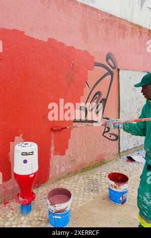 Processus d'un peintre en combinaisons vertes comme il peint sur des graffitis sur un mur rouge, couvrant de vieilles étiquettes avec de la peinture fraîche d'un rouleau Banque D'Images