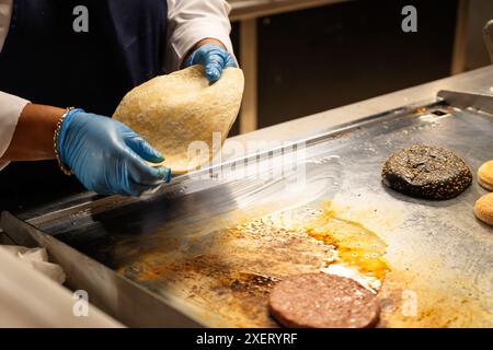 Une personne fait cuire des aliments sur un gril. La nourriture est un taco et un hamburger. La personne porte des gants Banque D'Images