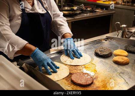 Une femme fait cuire des aliments sur un gril. Elle porte des gants et un tablier. Il existe plusieurs types d'aliments sur le gril, y compris les hamburgers et Banque D'Images