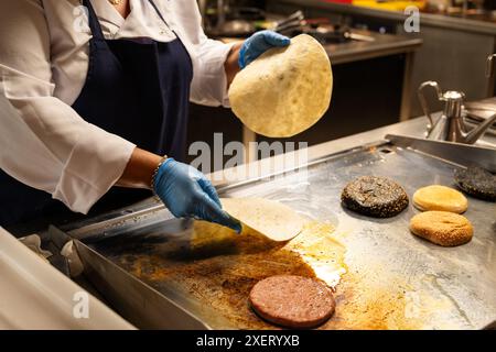 Une femme fait cuire des aliments sur un gril. Elle porte un tablier bleu et des gants. La nourriture sur le gril comprend des hamburgers et des tortillas Banque D'Images