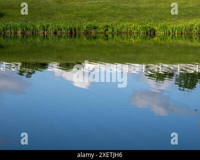 Dans un étang du quartier de Virginie, le ciel, les nuages et une clôture blanche sont magnifiquement reflétés dans l'eau. Banque D'Images