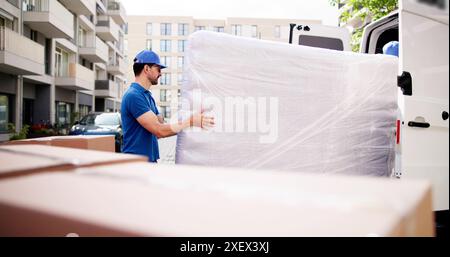 Déménageurs dédiés en action : hommes qui travaillent dur déchargeant le matelas enveloppé du camion, fournissant le service de livraison Top-Notch dans la rue animée de la ville. Banque D'Images