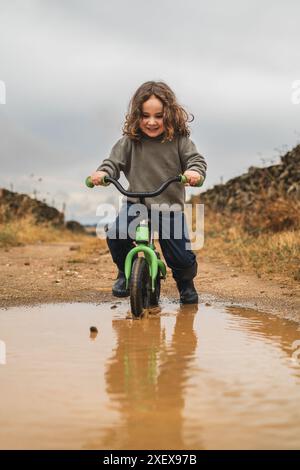 Une jeune fille roule sur un vélo vert à travers une flaque boueuse. La scène est ludique et légère, avec le sourire de la fille et la couleur vive du vélo a Banque D'Images