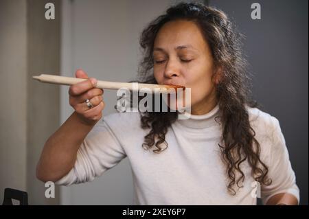 Portrait en gros plan d'une femme essayant de la sauce pour pâtes pour tester le goût. Jeune femme italienne jolie dégustation de nourriture, appréciant le goût, debout avec son œil Banque D'Images