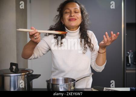 Belle femme italienne exprimant une émotion positive, dégustant la sauce tomate tout en cuisinant un plat dans la cuisine maison minimaliste moderne de couleur grise, express Banque D'Images