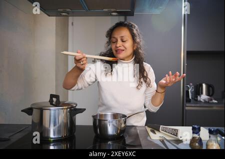 Femme préparant des spaghettis italiens selon la recette traditionnelle, testant la sauce pour pâtes à la tomate pour le goût, souriant à la caméra, debout à l'induc électrique Banque D'Images