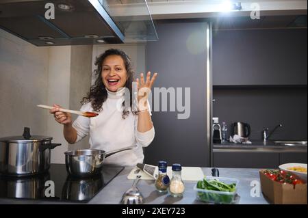 Jeune femme au foyer, femme de ménage, chef, cuisinier, femme latine faisant signe de délice de goût correct, savourant, préparant de la nourriture délicieuse, debout au comptoir de la cuisine Banque D'Images