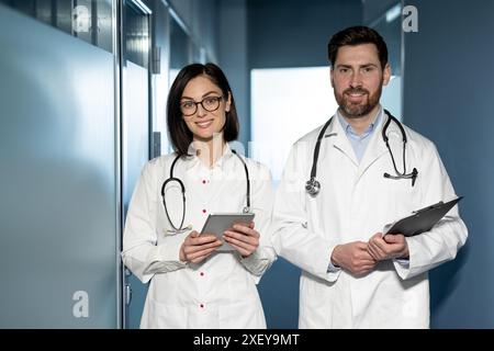 Professionnels de santé debout à l'hôpital tenant un comprimé et une planche à pince. Médecins portant des blouses de laboratoire blanches et des stéthoscopes. Travail d'équipe professionnel, service de santé, cadre hospitalier. Banque D'Images