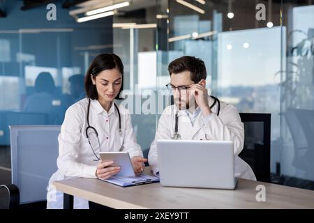 Professionnels de la santé en blouses de laboratoire collaborant dans un environnement de bureau moderne utilisant un ordinateur portable et une tablette pour la discussion et la planification. Collègues travaillant ensemble sur un projet de soins de santé Banque D'Images