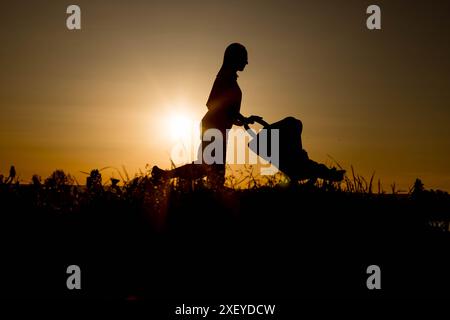 Silhouette d'une mère tirant une poussette contre le coucher du soleil près du lac. Banque D'Images