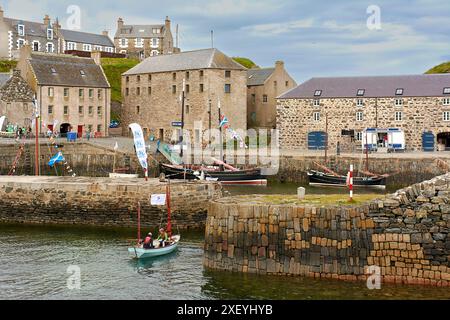 Portsoy Boat Festival un petit bateau à rames avec des voiles brunes et deux marins entrant dans le vieux port Banque D'Images