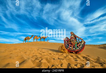 Une danseuse vibrante du Rajasthani tourbillonne sur les dunes de sable de Jaisalmer, avec une ligne de chameaux et leur berger en arrière-plan sous un drame bleu vif Banque D'Images