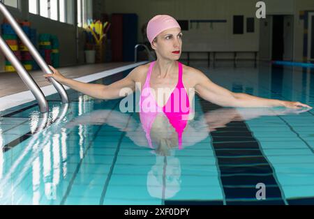 Femme d'âge moyen portant un maillot de bain rose et un bonnet de bain, se relaxant au bord d'une piscine intérieure, tenant sur l'échelle. Banque D'Images